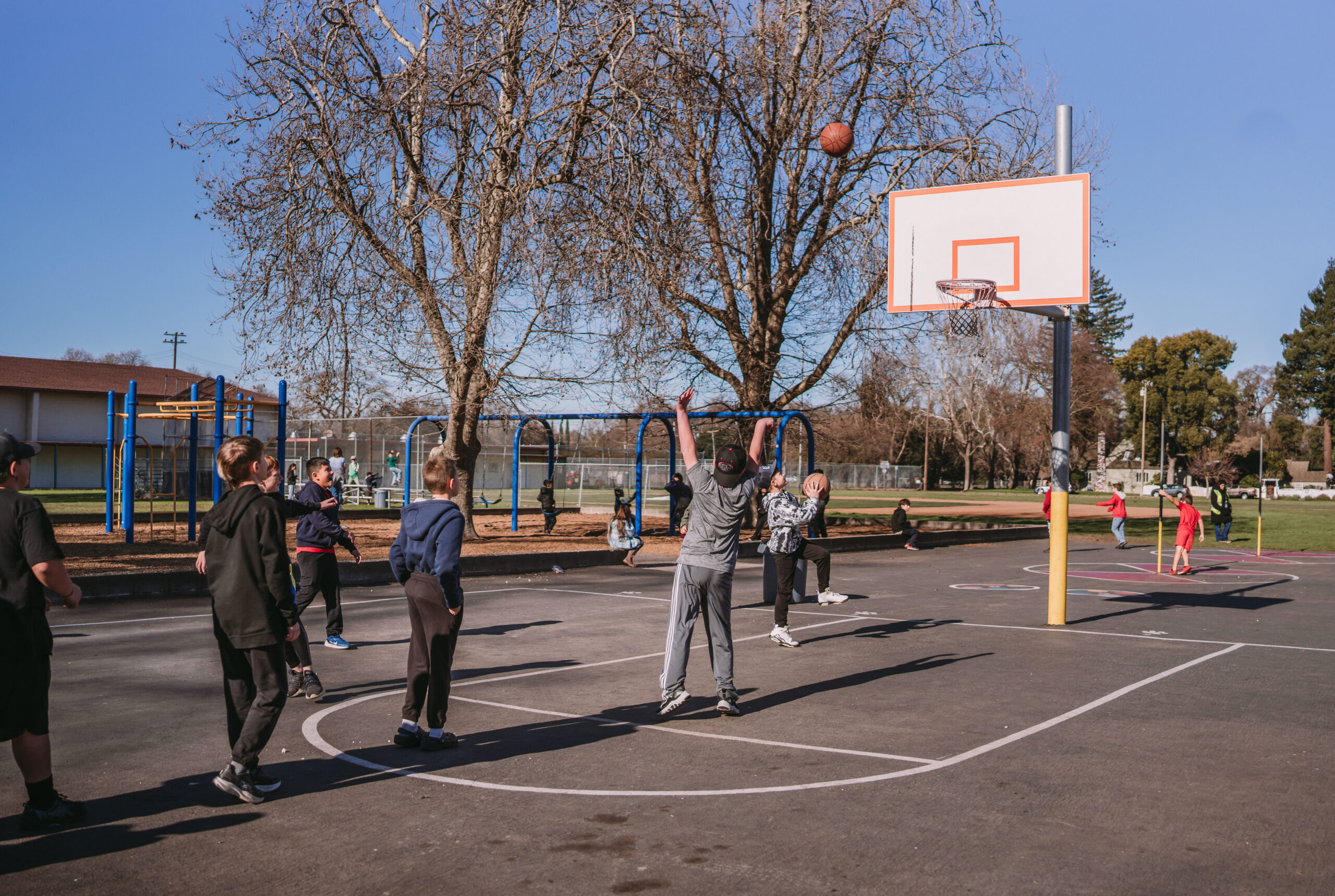 A group of boys laying basket ball watching one boy shoot a shot and waiting for it to go in.